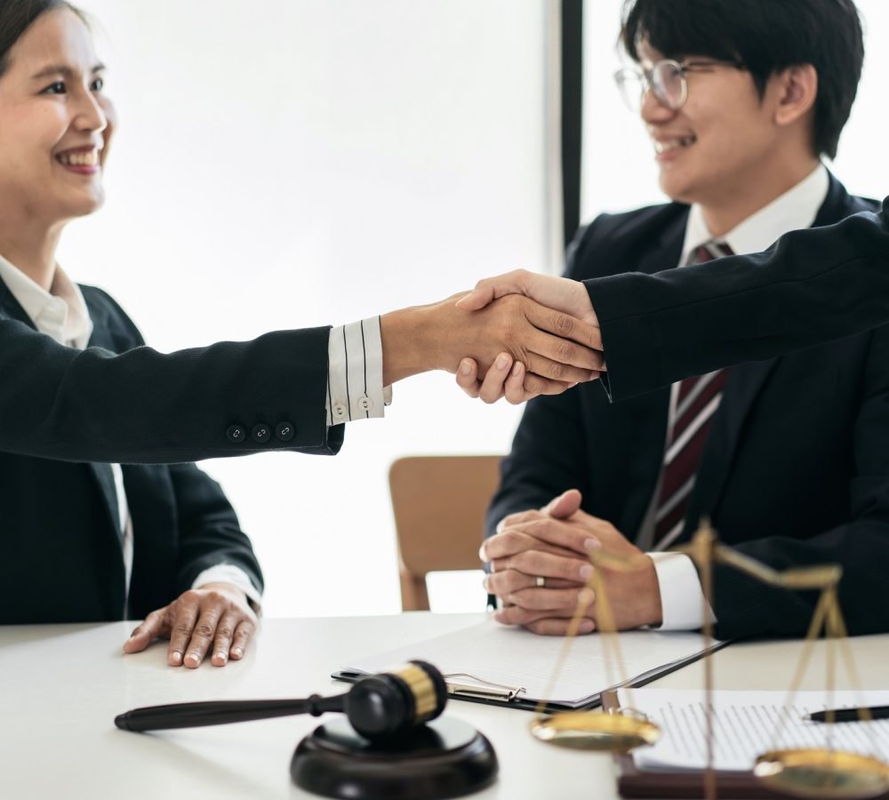 Female lawyer shaking hands with client after discussing deal of agreement together and signing contract on the table with brass scales and justice hammer in law firm.