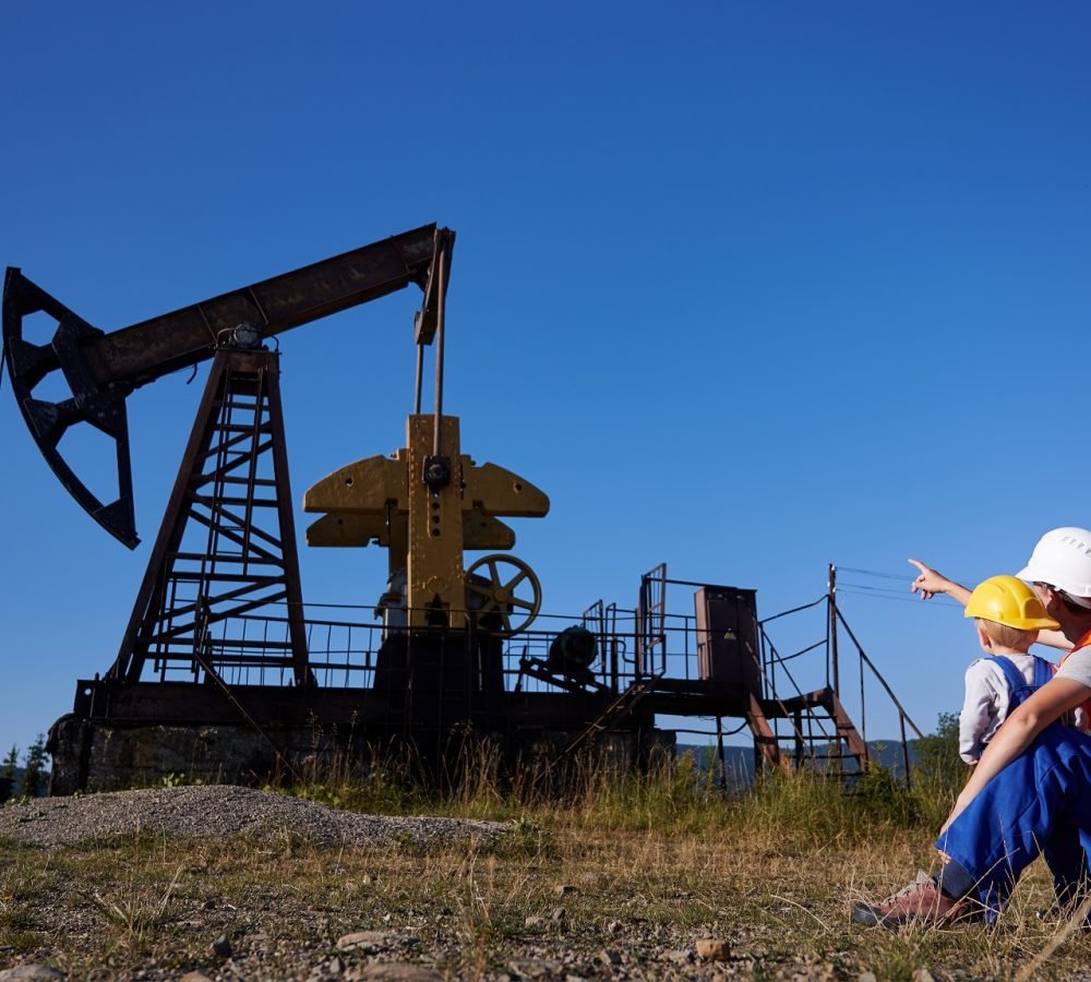 Back view of engineer pointing index finger at petroleum borehole pump in the oil field to preschool boy. Adult, acting mechanic and his little future successor observing the operation of oil pump.