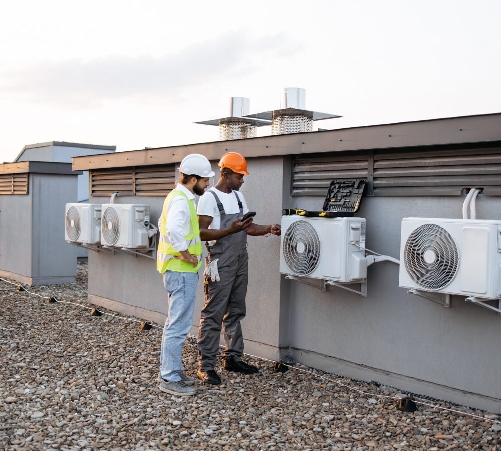 Group of multicultural construction workers on roof inspecting cooling system together using testing tool. Large and gray air conditioning unit with multiple ventilation openings and pipes.